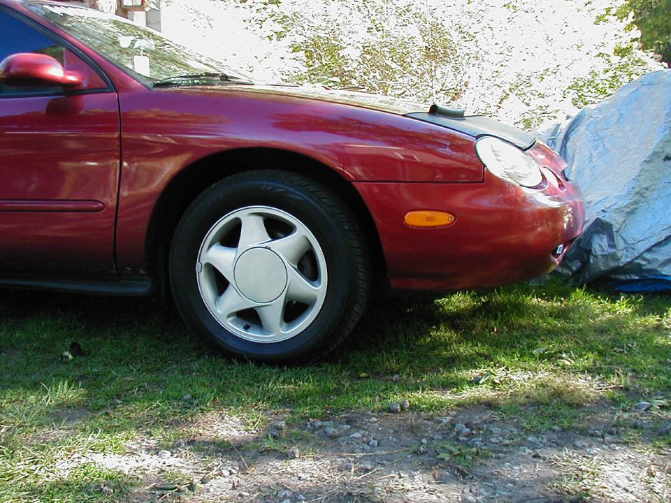 Dark red Ford Taurus front passenger side with alloy wheel and hood bra, parked on grass next to a tarp-covered object