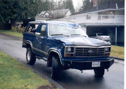 1989 Ford Bronco - This damaged blue Ford Bronco sits on a wet road, its roof and windshield severely compromised.