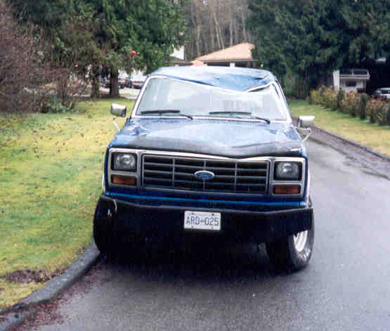 Blue Ford Bronco with dented roof, front view, parked on a wet street