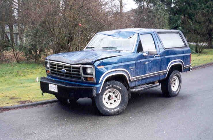 Blue Ford Bronco parked on a wet road, hood and windshield are damaged