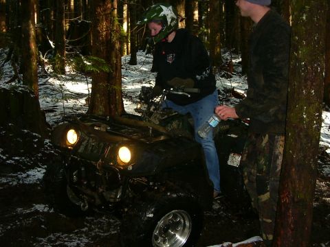 1990 Ford Bronco - Two individuals are on an all-terrain vehicle stuck in the mud and snow within a wooded area.
