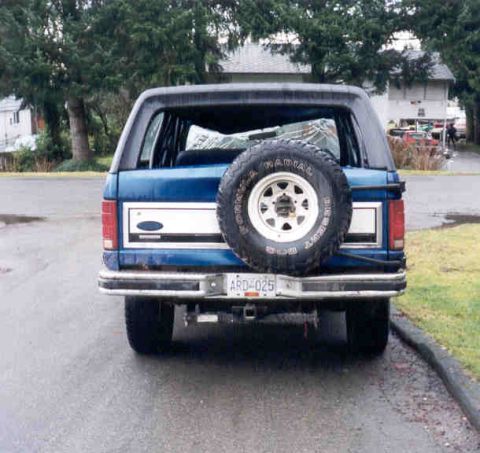 1989 Ford Bronco - This 1989 Ford Bronco is parked on a wet street with trees and houses in the background.