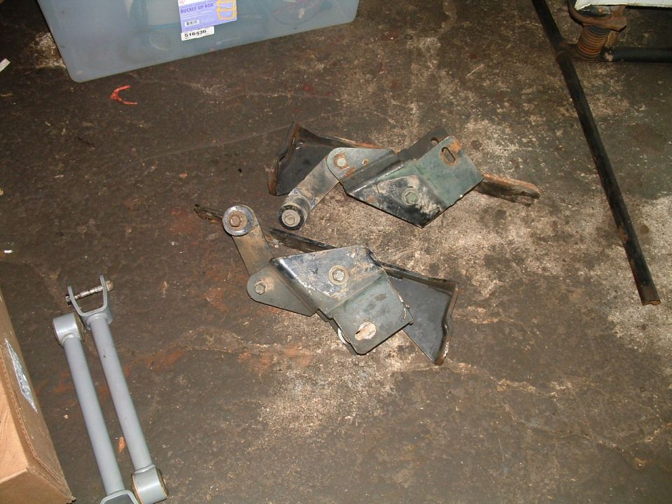 Two black metal brackets and two gray suspension arms on a dirty concrete floor
