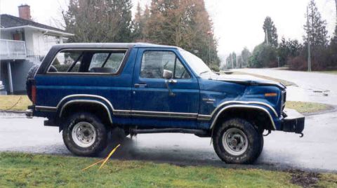 1989 Ford Bronco - Side view of a blue full-size Ford Bronco with a black hardtop, parked on wet pavement at a residential curb, showing collision damage that…
