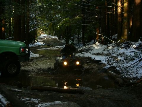 1990 Ford Bronco - A green 1990 Ford Bronco is parked on a snowy forest road while an ATV navigates through a muddy puddle.