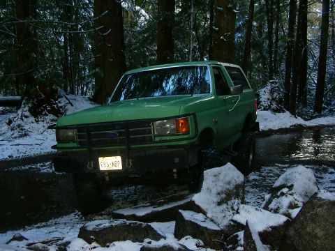 1990 Ford Bronco - Green 1990 Ford Bronco navigates a snowy, wooded trail with water and rocks.