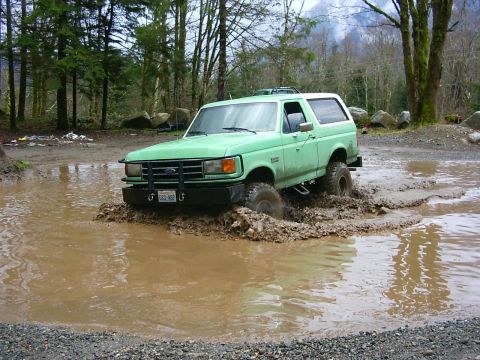 1990 Ford Bronco - A green 1987 Ford Bronco navigates a muddy obstacle course in a wooded area.