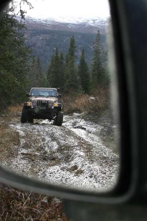 2003 Jeep TJ - A tan 2003 Jeep Wrangler navigates a challenging muddy and snowy trail through a forest, viewed from the side mirror.