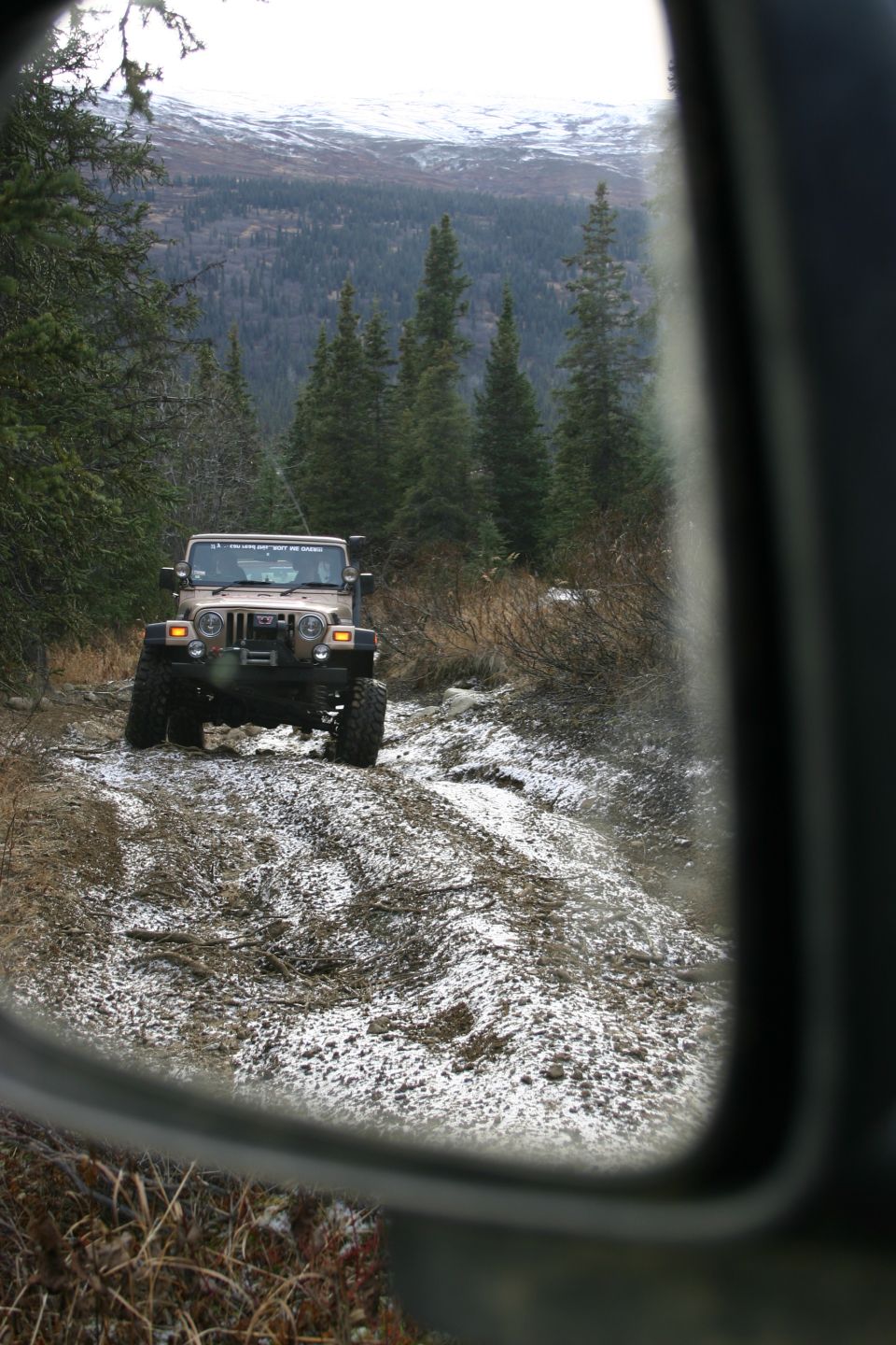 Tan 2003 Jeep Wrangler driving on a muddy, snowy trail, seen in side mirror