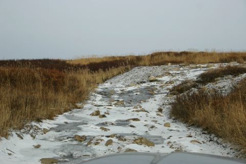 2003 Jeep TJ - A vehicle navigates a treacherous, icy, and rocky path through a landscape of dry grass under an overcast sky.