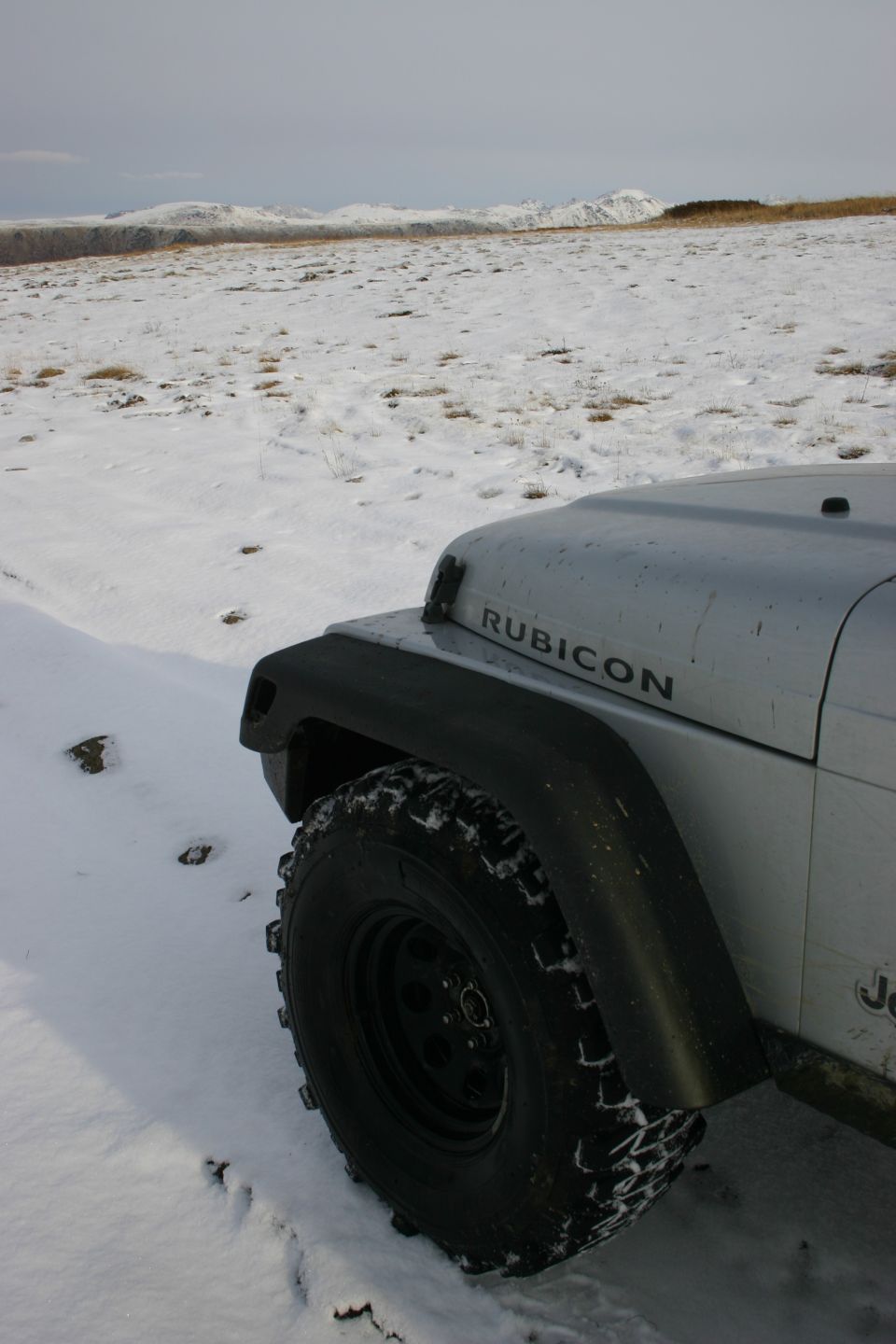Silver Jeep Wrangler Rubicon with aggressive tires in snow. 'RUBICON' text visible on hood