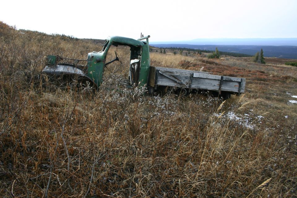 Old green truck sitting in a field of dry grass. It has a wooden flatbed and a missing hood