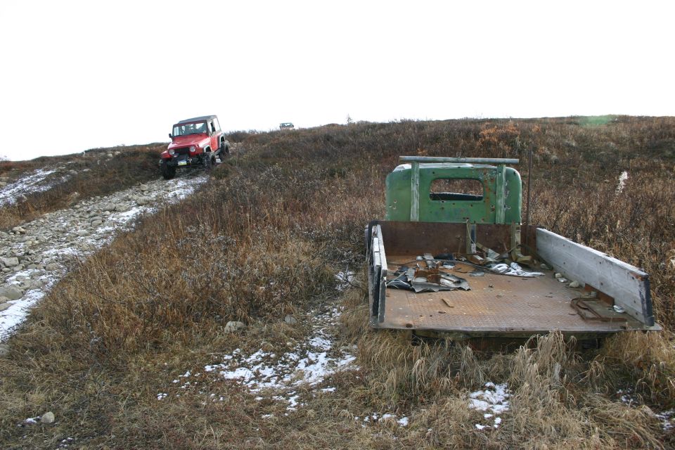 Red Jeep Wrangler driving on a rocky hill with a green flatbed truck in the foreground