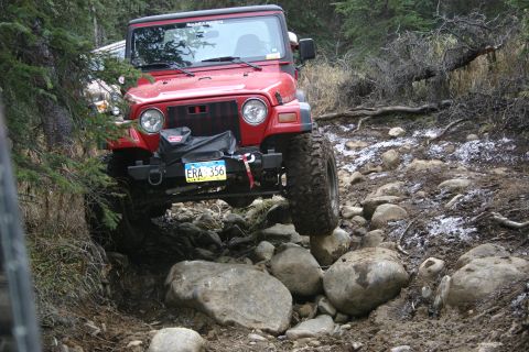 2003 Jeep TJ - A red 2003 Jeep TJ tackles a challenging, rocky off-road path with trees and debris surrounding it.