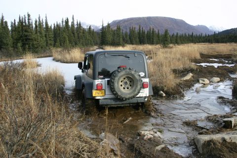 2003 Jeep TJ - A silver 2003 Jeep TJ navigates a challenging muddy and icy terrain with a wooded landscape and mountains in the background.