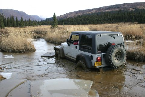 2003 Jeep TJ - A silver Jeep TJ navigates through a muddy, icy stream in a wooded, mountainous landscape.