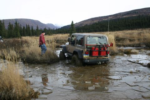 2003 Jeep TJ - A tan Jeep Wrangler TJ navigates a muddy, icy stream with two red jerry cans mounted on its rear carrier.