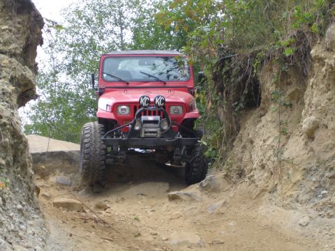 1990 Ford Bronco - A red Jeep Wrangler navigates a narrow, rocky off-road trail with steep dirt banks on either side.