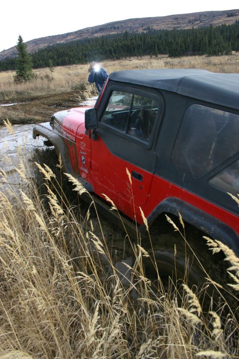 2003 Jeep TJ - A red 2003 Jeep TJ Wrangler is partially submerged in mud and tall grass, with a person standing near the hood.