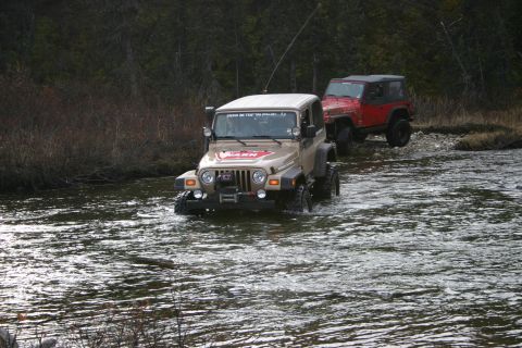2003 Jeep TJ - Two Jeep Wranglers navigate a river crossing, one tan and one red, with trees in the background.
