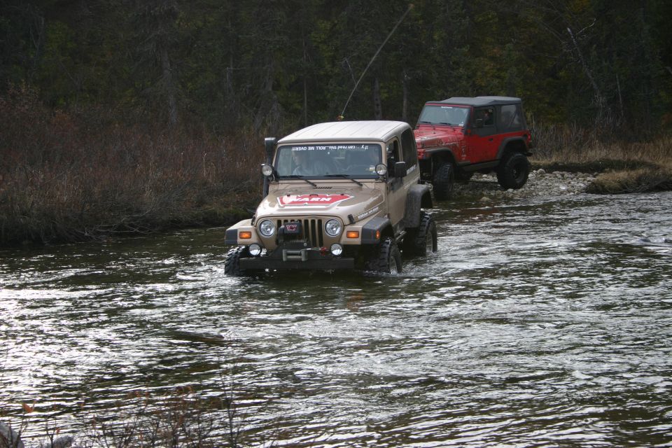 Two Jeep Wranglers, one tan and one red, driving through a river. Tan Jeep has a WARN winch and a CB antenna