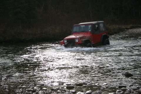 2003 Jeep TJ - A red Jeep TJ navigates a shallow, rocky riverbed, kicking up water as it goes.