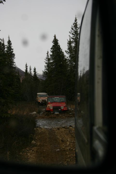 2003 Jeep TJ - Two Jeeps navigate a muddy trail through a forest with snow-capped mountains in the distance.