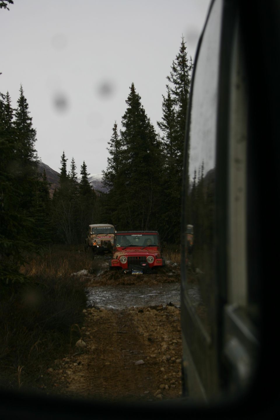 Red Jeep TJ and tan Jeep Wrangler driving through mud on a trail. The red Jeep is in the foreground