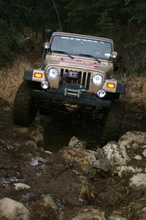 2003 Jeep TJ - A tan Jeep Wrangler TJ tackles a challenging off-road obstacle on a wooded trail.