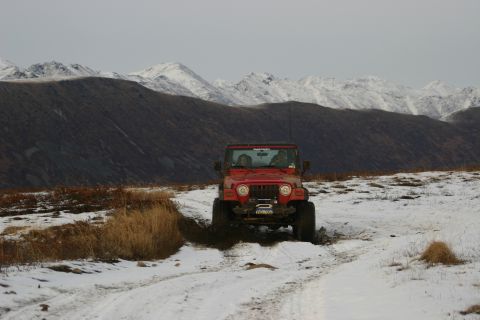 2003 Jeep TJ - A red 2003 Jeep TJ navigates a muddy, snow-dusted path with mountains in the background.