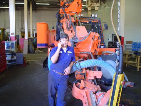1966 International School Bus - A worker stands in a garage, talking on his phone next to a large piece of orange industrial machinery.