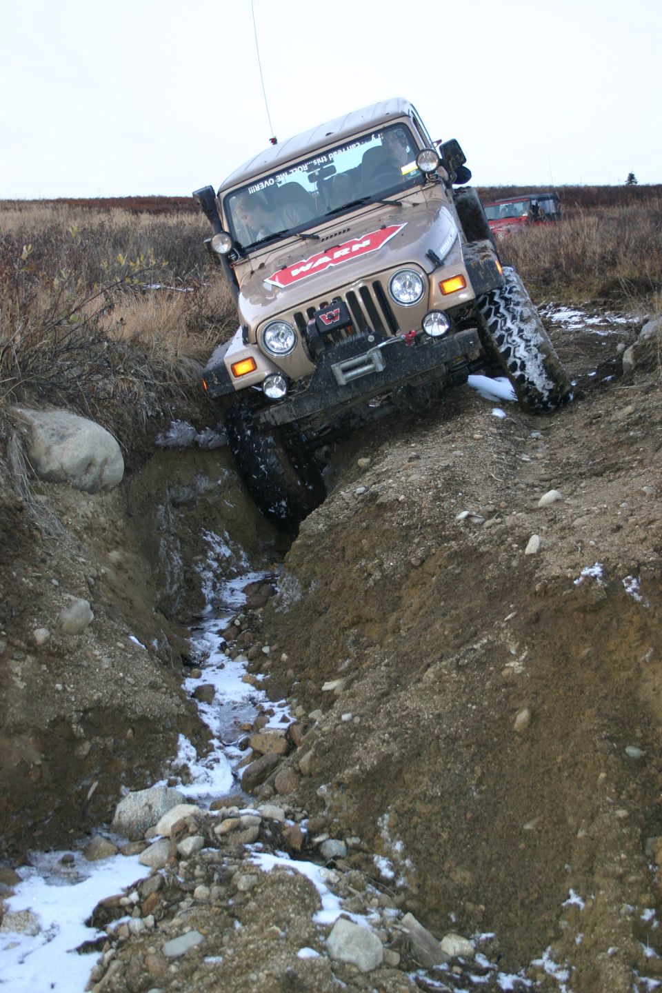 Tan 2003 Jeep TJ driving through a muddy ditch with snow patches