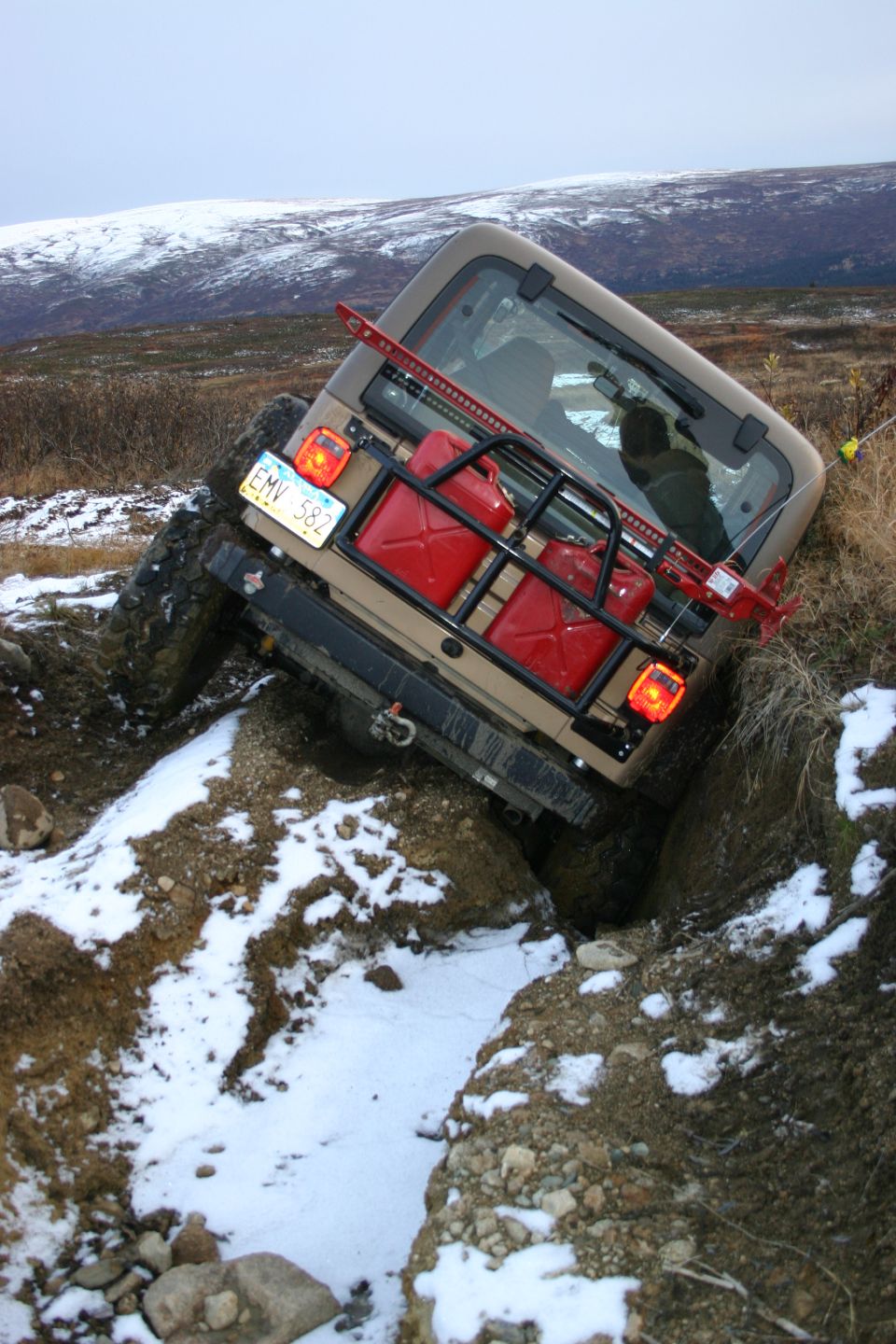 Tan Jeep TJ with two red gas cans on rear rack, stuck in a ditch with snow on the ground