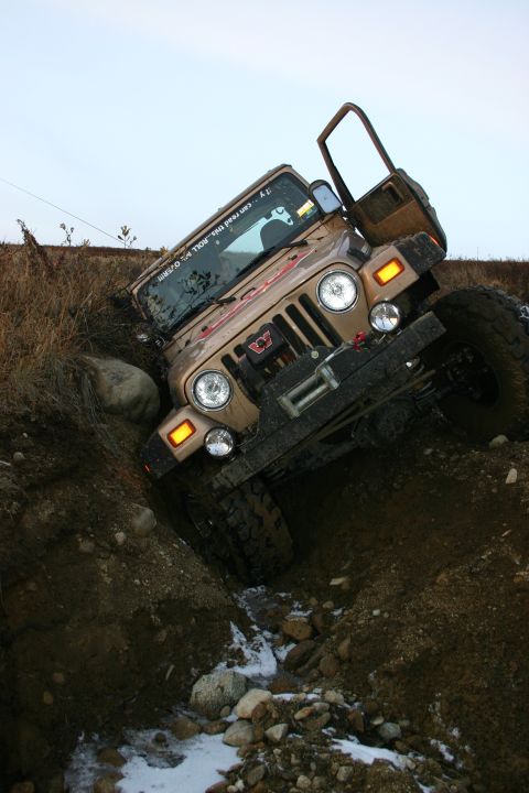 2003 Jeep TJ - A tan Jeep TJ is tilted precariously in a muddy ditch with snow patches on the ground.