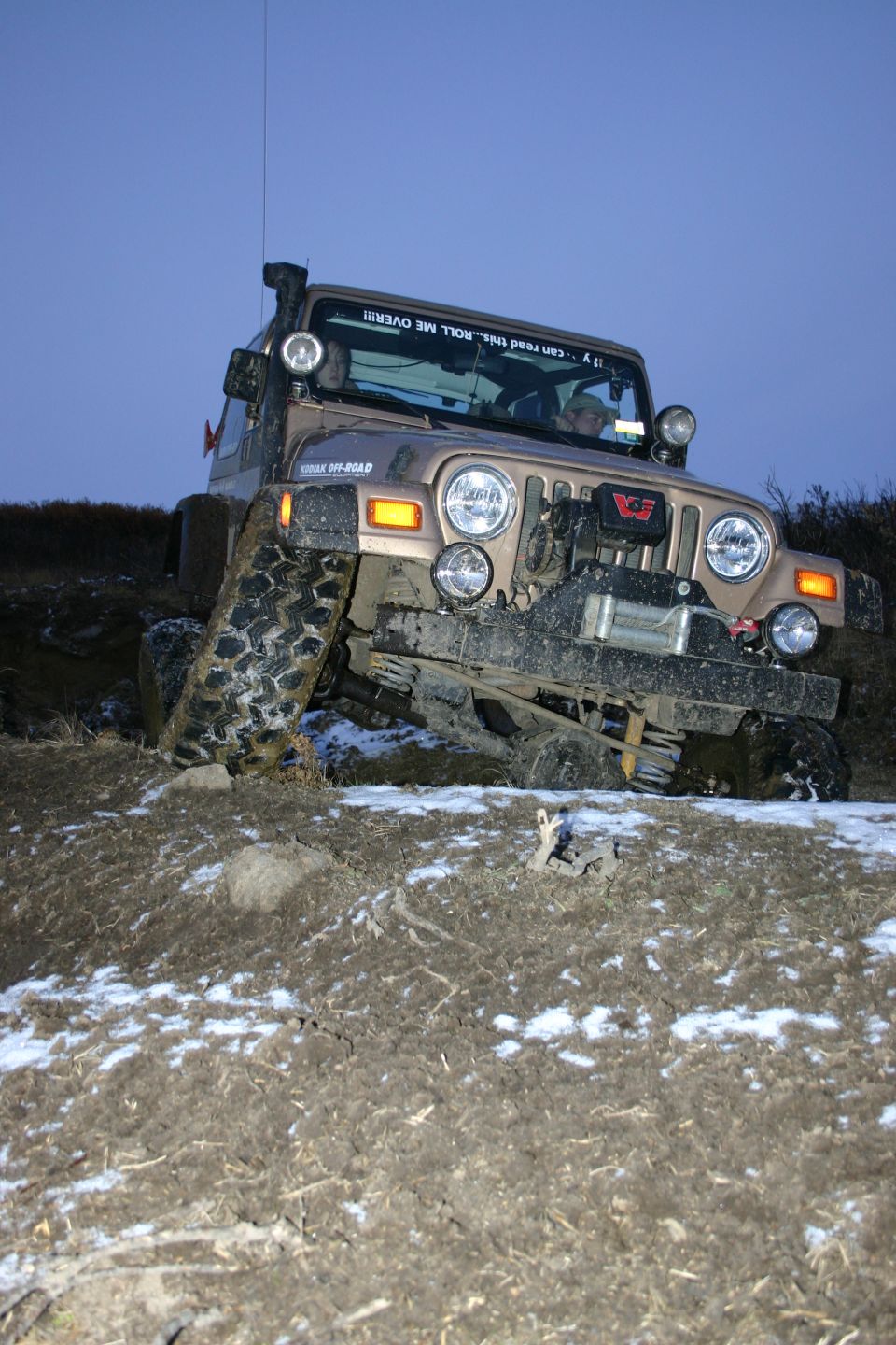 Tan 2003 Jeep TJ off-roading on a muddy slope with snow patches, front 3/4 view