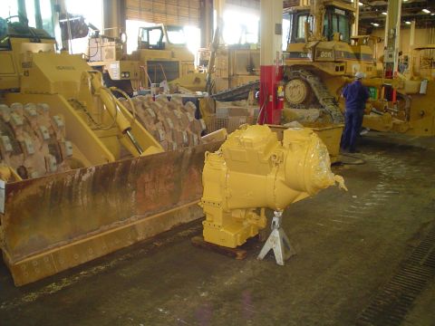 1966 International School Bus - A yellow transmission is displayed on jack stands in a workshop, with various pieces of heavy machinery visible in the background.
