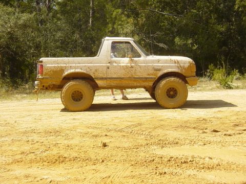 1989 Ford Bronco - Mud-covered 1989 Ford Bronco on a dirt track with trees in the background.
