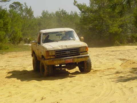 1989 Ford Bronco - A lifted Ford F-150 navigates a sandy, off-road trail with trees in the background.