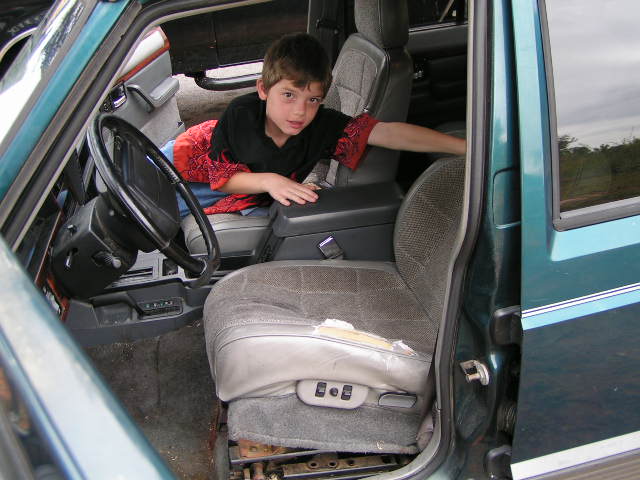Boy leaning between gray cloth front seats inside a teal SUV, seen through open driver's door