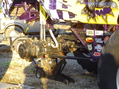 1979 Ford Bronco - Close-up of a monster truck's rear axle assembly being worked on, with a smaller vehicle behind it.