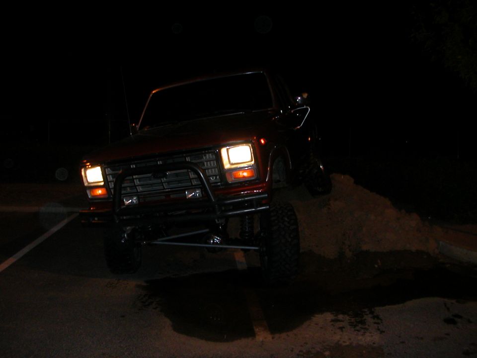 Red Ford Bronco with bull bar, headlights on, climbing a dirt pile at night