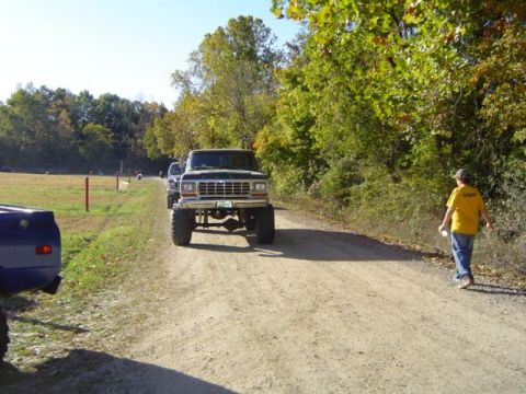 1979 Ford Bronco - A lifted pickup truck drives down a dirt road with trees and a field in the background.