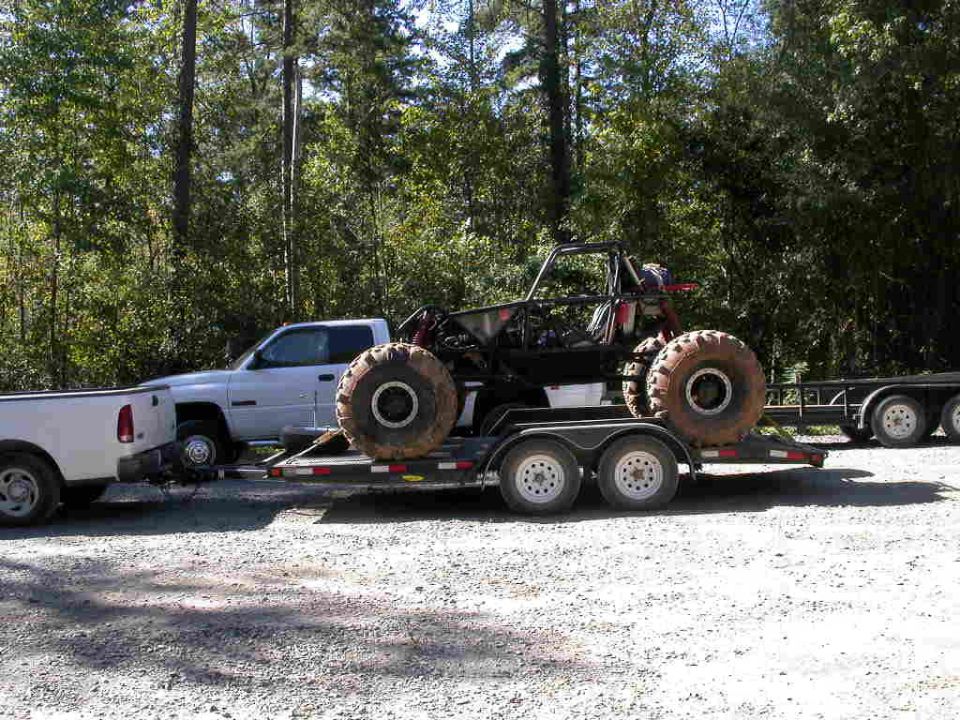 Tube-frame buggy with large mud tires loaded on a flatbed trailer behind a white Dodge dually