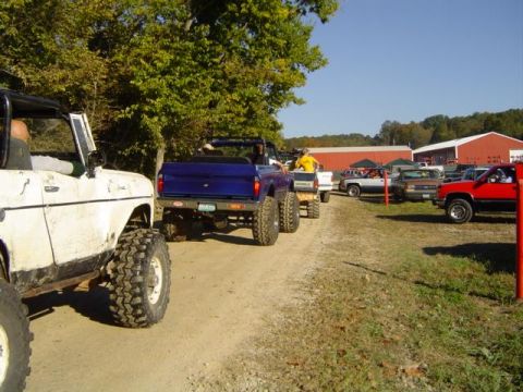 1979 Ford Bronco - Several off-road vehicles are gathered on a dirt path near buildings and trees under a clear blue sky.