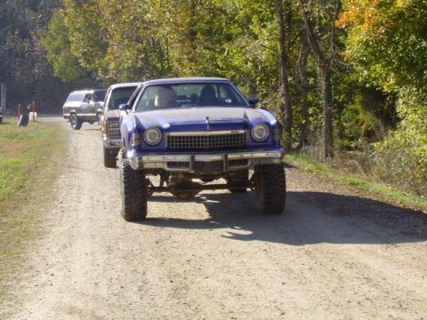 1979 Ford Bronco - A line of lifted vehicles, including a blue 1975 Chevrolet Monte Carlo, drive down a dirt road.