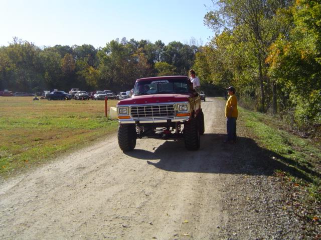 Red 1979 Ford Bronco lifted on a dirt road with people standing nearby. Other vehicles parked in the background