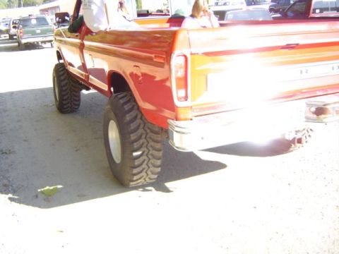 1979 Ford Bronco - A lifted red pickup truck with oversized tires is parked on a dirt lot, with other vehicles in the background.