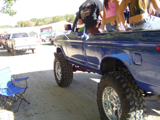 Blue 1979 Ford Bronco lifted with large tires, people sitting in the bed. Another white pickup truck is parked nearby