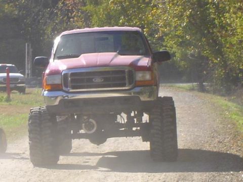 1979 Ford Bronco - A lifted Ford truck kicks up dust on a rural road, with another vehicle visible in the background.