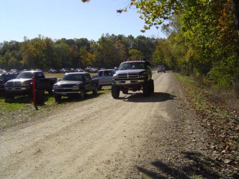 1979 Ford Bronco - Several lifted pickup trucks are seen on a dirt road during an outdoor event, with more vehicles parked in the background.